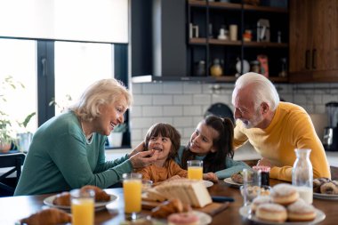 İki mutlu küçük çocuk, erkek ve kız büyük anne ve babasıyla kahvaltı yapıyor. Gülümseyen güzel yaşlı kadın büyükannesi evde kahvaltıda küçük tatlı torununu besliyor..