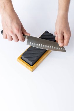 Masanın beyaz arka planında, erkek elleri kesiklere karşı çelik bıçak korumasını keskinleştiriyor, bıçakları bilemek için orta sert siyah bir taş profesyonel bıçakları kesmek için keskinleştiriyor.