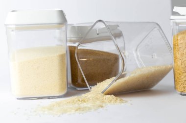 Yakın plan beyaz, üç farklı boyutta şeffaf konteynır var. İçi pirinç ve kahverengi şeker ile doldurulmuş mısır gevreğiyle dolu beyaz kapaklar ve bir konteynırdan dökülen pirinç.