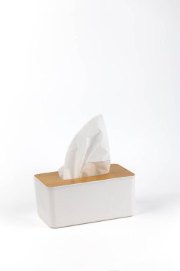 Beyaz arka planda beyaz bir plastik peçete tutacağı duruyor. Tahta kapaklı, minimalist bir tasarımı var. Beyaz peçete hijyen setini dışarı çıkarıyor.