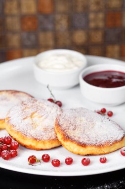 Siyah masaya yakın çekim. Kızarmış peynirli kekler, ekşi krema ve kırmızı sonbahar meyveli çilek reçeli.