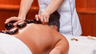 Masaj salonuna yakın çekim, iyi ışıklandırma, müşteri masanın üzerinde yatıyor, masör siyah cepli, beyaz üniformalı ve havluyla taş masajı yapıyor, kas gevşetici.