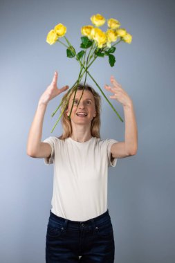Açık gri bir arka planda açık renk tişörtlü genç bir kız yüzünde ayrılık gülümsemesi için 8 Mart 'taki sarı çiçeklerin önüne şakayık güller atıyor.