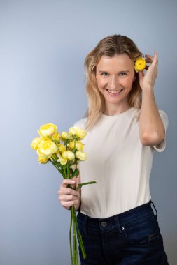 Açık gri arka planda açık renk tişörtlü genç bir kız elinde sarı şakayık gülleri 8 Mart gülümsemesi saçlarında tomurcuklar, kameraya bakıyor.