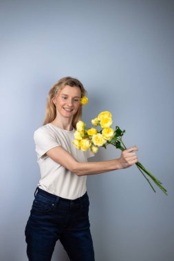 Açık gri bir arka planda açık renk tişörtlü genç bir kız yüzünde ayrılık gülümsemesi için 8 Mart 'taki sarı çiçeklerin önüne şakayık güller atıyor.