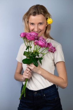 Açık gri arka planda açık renk tişörtlü genç bir kız elinde pembe şakayık güller 8 Mart gülümsemesi saçlarında tomurcuklara kameraya bakıyor
