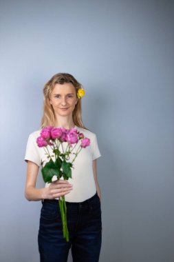 Açık gri arka planda açık renk tişörtlü genç bir kız elinde pembe şakayık güller 8 Mart gülümsemesi saçlarında tomurcuklara kameraya bakıyor