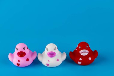 Pembe, beyaz ve kırmızı renkli üç sevimli plastik ördek, kalp desenleriyle süslenmiş, canlı mavi bir arkaplanda oturuyor, her yere neşe ve neşe katıyor..