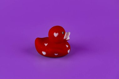 Bu resim, canlı mor bir arkaplan, banyo dekoru ve çocuk oyuncakları için ideal, beyaz kalp desenleriyle süslenmiş kırmızı kauçuk bir ördeği gösteriyor..