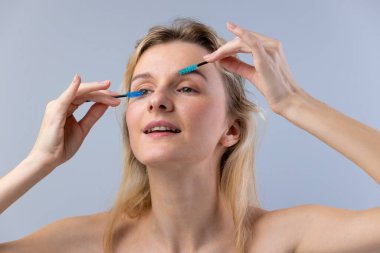 Görüntü, kirpiklerini iki renkli fırçayla tarayan, gülümseyen ve güzellik rutininin tadını çıkaran bir kadını gösteriyor. Basit, gri bir arkaplana yaslanmış, doğal görünüşünü vurgulayan bir kadın..