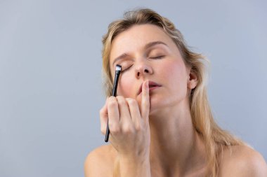 This image showcases a tranquil woman focusing on her makeup application. She uses a brush to apply eyeshadow delicately, emphasizing her natural beauty in a subtle, artistic manner. clipart