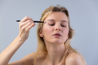 Bu resimde, odaklanmış genç bir kadın bir fırçayla göz kapağına nazikçe makyaj yapıyor, doğal güzelliğini ve kişisel bakım ve kendini ifade etmenin önemini vurguluyor..