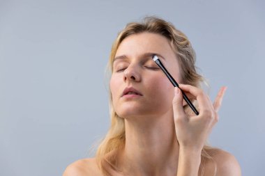 Bu görüntü, makyajı fırçayla yaparken gözleri kapalı sakin bir kadını yakalar. İfadesi kendine güven ve sükunet yayıyor, özgüven ve güzellik sanatını gözler önüne seriyor..