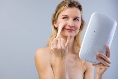 Bu resimde, genç bir kadının el aynasına bakarken fırça kullanarak allık sürdüğü görülüyor. Dekor güzellik, özgüven ve kişisel bakım konularını vurguluyor.