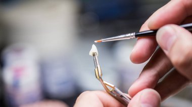 Bir diş teknisyeninin, bir laboratuvar ortamındaki dişçilik sürecinde ince bir fırça kullanarak dişçi kaplamasını titizlikle elle boyamasına odaklanın..