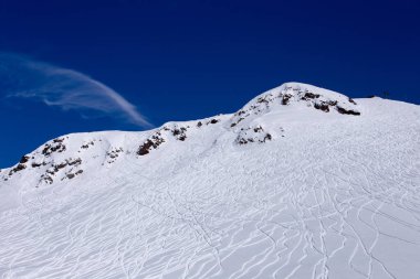Bu resim, temiz mavi gökyüzü ile karla kaplı, el değmemiş kar ve kayalık araziye oyulmuş çeşitli kayak yollarını gösteren çarpıcı bir dağ manzarası yakalıyor..