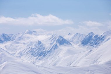 Güneşin altında parlayan görkemli tepeleri gösteren karlı bir sıradağ manzarası yumuşak bulutlarla birlikte, kış manzaralarının ve doğanın güzelliğini gösteriyor..