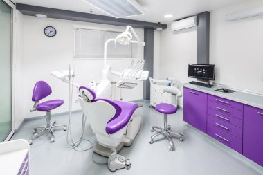 Bu resimde mor sandalyeler, gelişmiş diş aletleri ve profesyonel ve misafirperver bir çevreyi yansıtan temiz ve parlak bir çalışma alanı ile donatılmış modern bir diş muayenehanesi sergilenmektedir..
