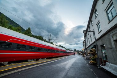 Bu resim Norveç istasyonunda hareket eden canlı bir kırmızı treni gösteriyor. Bulutlar ve ilginç bir sanat eseriyle tamamlanarak benzersiz bir atmosfer yaratıyor..
