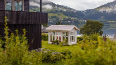 Bu resimde, geniş pencereli, canlı yeşilliklerle çevrili, dağların arkasına kurulmuş güzel bir bahçe evi ve huzur dolu bir göl var..