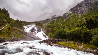 Bu çarpıcı görüntü kayalık arazide çağlayan güçlü bir şelale yakalıyor, yeşil ormanlar ve yüksek dağlarla çevrili, sükunet ve doğa güzelliğini çağrıştırıyor..