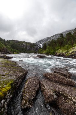 Bu resim dokulu kayalar arasında akan sakin bir nehri yakalar, muhteşem bir şelale manzarası ve arka plandaki dağlar, hepsi nazik bulutlarla kaplıdır..