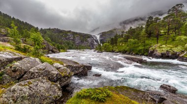 Bu çarpıcı görüntü kayaların üzerinde süzülen sakin bir nehri yakalıyor, etrafını saran yeşil ağaçlar ve bulutlarla örtülü dağlar, nefes kesici bir doğal ortam yaratıyor..