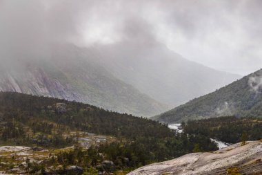 Bereketli yeşillik ve dolambaçlı bir nehir ile kaplanmış Norveç engebeli dağlarının dingin güzelliğini deneyimleyin. Bu el değmemiş doğal manzaranın nefes kesici cazibesini ekleyin..