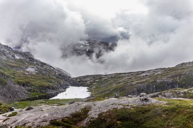 Dramatik bulutlar, kayalık arazi ve yemyeşil verimli doğal ortam yaratan sakin bir gölün yer aldığı Norveç dağlarının hayranlık uyandırıcı manzarası..