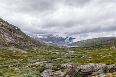 Bu çarpıcı görüntü, Norveç 'in yeşil tepeleri, kayalık çıkıntıları ve dramatik bulutların altındaki dingin çevreye yerleşmiş büyüleyici kırmızı bir kulübesini sergiliyor..