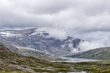 Norveç 'in engebeli engebeli arazisinin huzur dolu özünü keşfedin, yüksek dağların arasına yerleşmiş dingin göller barındırın. Karamsar bulutlar manzarayı güzelleştiriyor..