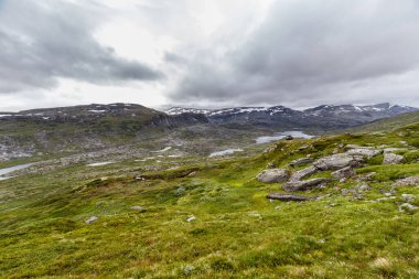 Büyüleyici İskandinav manzarasının geniş bir görüntüsü engebeli araziler, yumuşak tepeler ve bulutlu bir atmosferde karla kaplı dağlarla çevrili dingin göller..