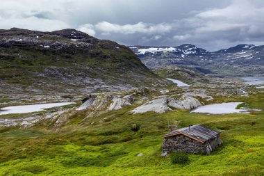 Bu çarpıcı görüntü, sakin göller ve yüksek dağlarla yeşil tepeler arasında kurulmuş uzak taştan bir kulübeyi sergiliyor ve Norveç 'in güzel çöllerinde sakin bir atmosfer yaratıyor..