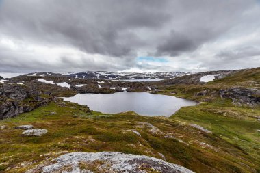 Bu nefes kesici manzara, yeşil tepeler ve kayalık oluşumlar tarafından kucaklanan sakin bir gölü sergiliyor. Hepsi de çarpıcı bulutlu bir gökyüzünün altında..
