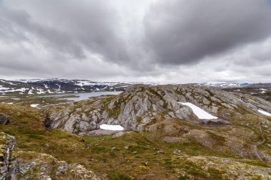 Norveç 'in çarpıcı manzarası engebeli kayalık tepeleri, karları ve kasvetli gökyüzünün altında sakin bir gölü sergiliyor, doğanın vahşi ve el değmemiş güzelliğini somutlaştırıyor..