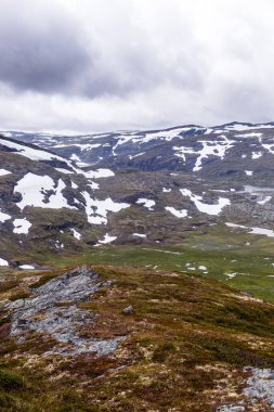 Canlı yeşilliklerle bezenmiş, karla kaplı tepeleri ve doğanın güzelliğini gösteren atmosferik bulutlu gökyüzünü keşfedin..