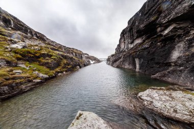 Sarp kayalıkların ve canlı yeşil yosunların sardığı sakin bir su yolu bulutlu bir gökyüzüne karşı kurulmuş, keşif ve merakı davet eden pitoresk bir doğal manzara yaratıyor..