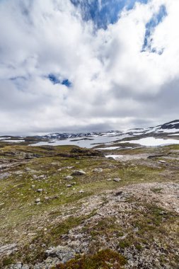 Bu görüntü, ön planda kayalık arazi ve arka planda dramatik bulutlar ve karla kaplı dağlarla çerçevelenmiş sakin bir göl içeren nefes kesici bir İskandinav manzarası yakalar..