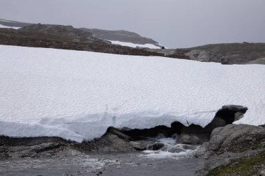 Bu resim, kalın buz tabakasının altında akan suyla karla kaplı çarpıcı bir manzara tasvir ediyor. Dağlık bir bölgedeki doğanın huzur dolu güzelliğini gözler önüne seriyor..