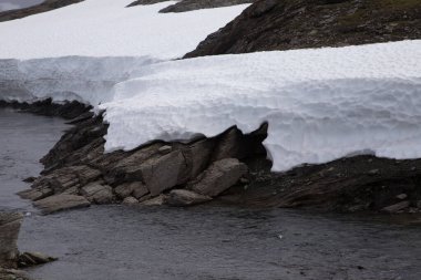 Bu büyüleyici görüntü, parlak beyaz kar örtülerinin bir nehrin akan sularıyla pürüzsüz bir şekilde birleşerek sakin ve manzaralı bir manzara oluşturduğu güzel bir karlı manzarayı gözler önüne seriyor..