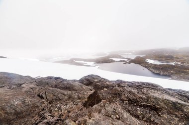 Bu büyüleyici görüntü, sisle kaplanmış sakin, karla kaplı bir araziyi ortaya çıkarıyor. Güzel kaya oluşumları ve durgun su vücutları sergiliyor..