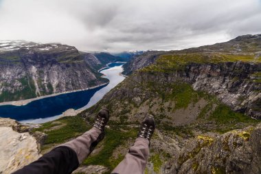 Cesur bir yürüyüşçü muhteşem bir fiyordun üzerinde uçurumun kenarında oturmanın heyecanını yaşar. Sahne, gri bulutlar altında dağlık araziler arasında salınan sakin bir nehri gösteriyor..