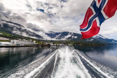 Norveç fiyordunu gösteren nefes kesici bir sahne görünür bir tekne uyanışıyla, dalgalanan bir Norveç bayrağı ve kısmen bulutlarla örtülü resimli dağlar..