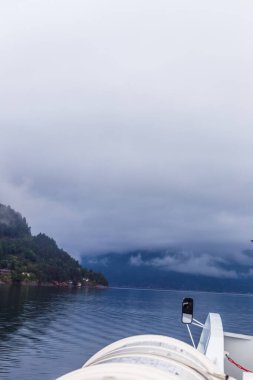 Bu görüntü, bir tekne, sis kaplı dağlar ve bulutlu gökyüzünü yansıtan sakin sularla huzurlu bir fiyort görüntüsü yakalar ve Norveç 'te doğal güzellik ortamı yaratır..