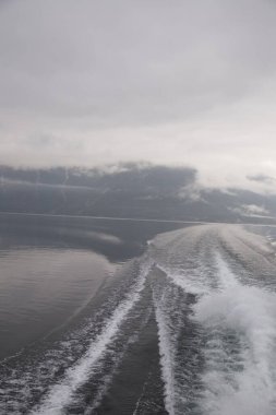 Bu görüntü, bulutlu bir gökyüzünün ve ufuktaki uzak dağların altındaki dingin göl manzarasını gösteren bir tekne tarafından oluşturulan sisli suların ve yumuşak dalgaların güzelliğini yakalar..
