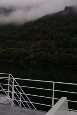 Feribottan çekilen sakin bir sahne bulutlarla gizlenmiş sisli dağları gözler önüne seriyor. Bereketli yeşil ormanlar sakin suları kaplıyor ve doğada barışçıl bir atmosfer yaratıyor..