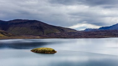 Bu çarpıcı görüntü durgun suların yemyeşil ile buluştuğu karamsar, bulutlarla dolu gökyüzünün altında sakin atmosferi yükselten görkemli dağlarla çevrili sakin bir manzara yakalıyor..