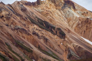 Çarpıcı renkler ve jeolojik oluşumlar gösteren sıradışı bir dağ manzarası. Kayalık dokular yeşil ve kar tarafından çerçevelenmiş minerallerin ve doğal renklerin katmanlarını ortaya çıkarıyor..