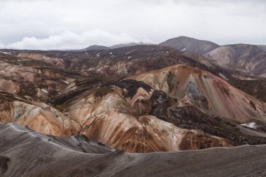 Görüntü, İzlanda 'daki volkanik aktivite tarafından şekillendirilen çok renkli dağların ve vadilerin geniş bir görüntüsünü sunuyor. Bulutlarla dolu dramatik bir gökyüzünün ortasındaki saf güzelliği yansıtıyor..