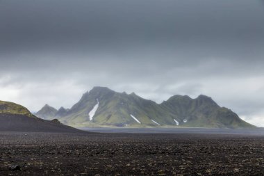 Bu çarpıcı görüntü, İzlanda 'nın eşsiz arazisinin büyüleyici güzelliğini, engebeli dağları ve dramatik, atmosferik gökyüzünün altında çıplak ve çorak bir manzarayı yakalar..